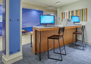 A reception desk with two monitors, a printer, and a keyboard. There are two chairs and a small table with a bench. The room has a blue accent wall and a carpeted floor. - Holiday Inn Express & Suites Hermiston Downtown by IHG (Hermiston)