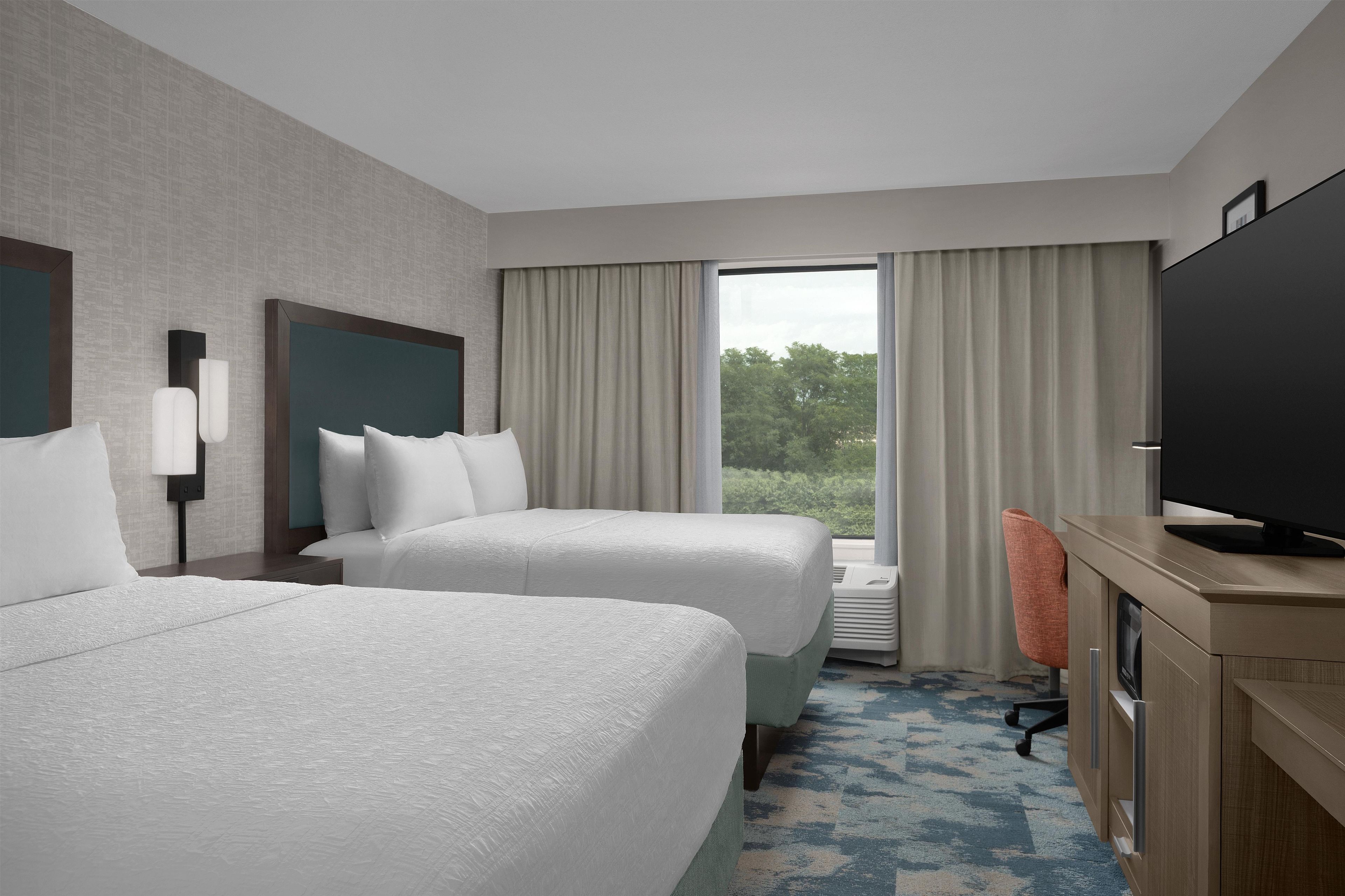 Une chambre d’hôtel avec deux lits, un bureau doté d’un téléviseur à écran plat et une grande fenêtre avec des rideaux.