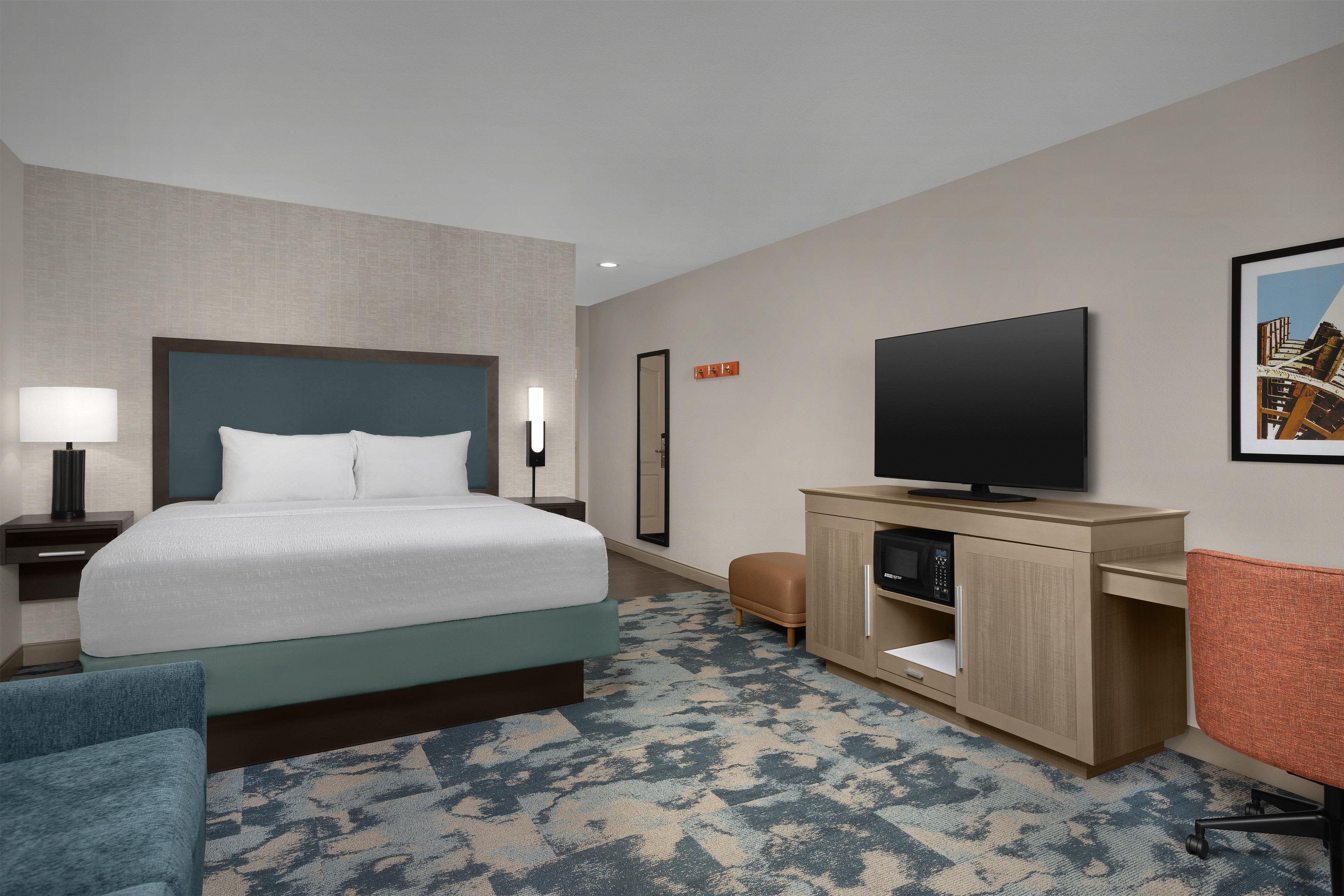 A hotel room with a bed, a desk with a flat-screen TV, a chair, a nightstand, and a wall-mounted TV.
