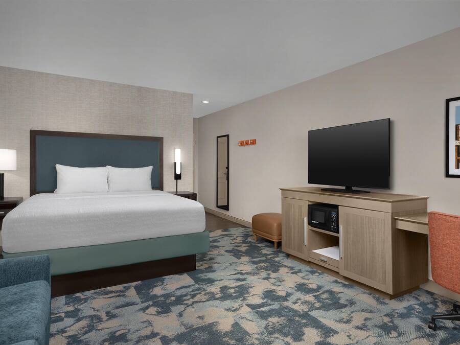 A hotel room with a bed, a desk with a flat-screen TV, a chair, a nightstand, and a wall-mounted TV.