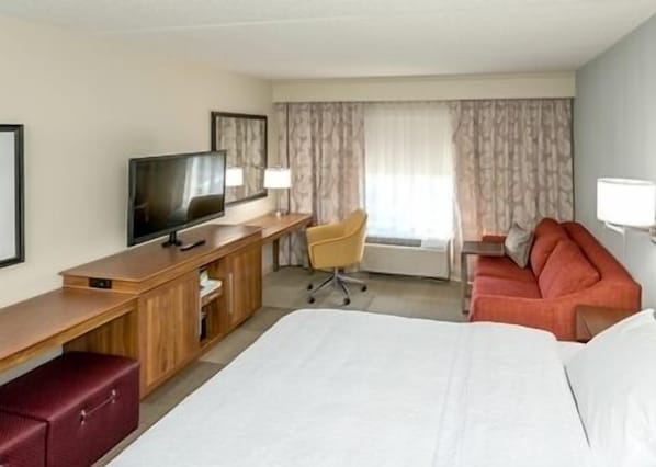 Quarto de hotel com cama, escrivaninha, televisão, poltrona, sofá e janela com cortinas.