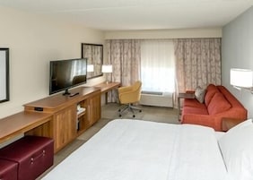 Habitación de hotel con cama, escritorio con televisor, silla, sofá y ventana con cortinas.