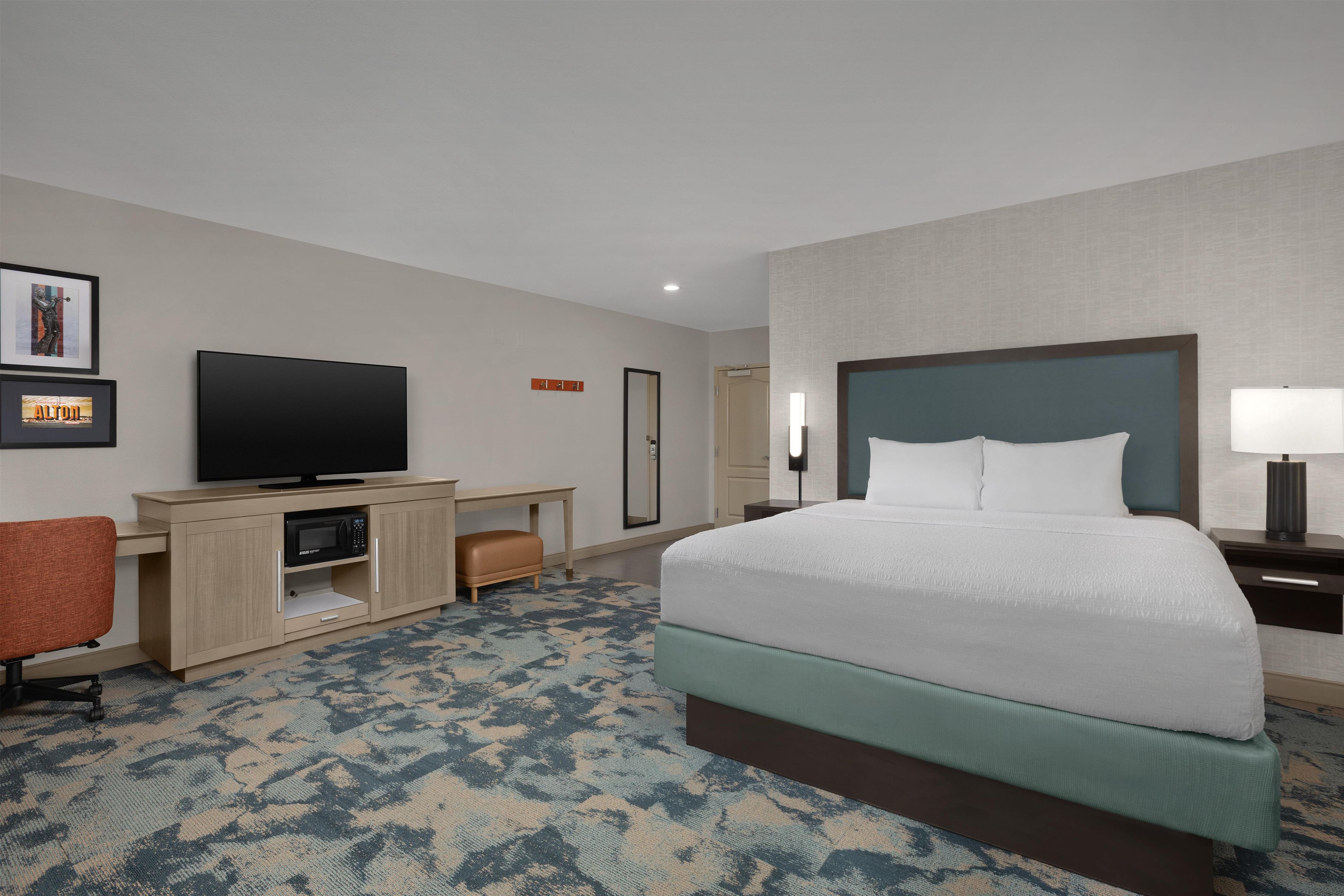 Een hotelkamer met een bed, een bureau met een flatscreen televisie, een stoel, een nachtkastje met een lamp en een spiegel.
