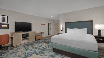 Een hotelkamer met een bed, een bureau met een flatscreen televisie, een stoel, een nachtkastje met een lamp en een spiegel.