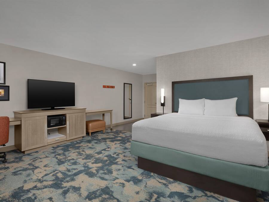 Een hotelkamer met een bed, een bureau met een flatscreen televisie, een stoel, een nachtkastje met een lamp en een spiegel.