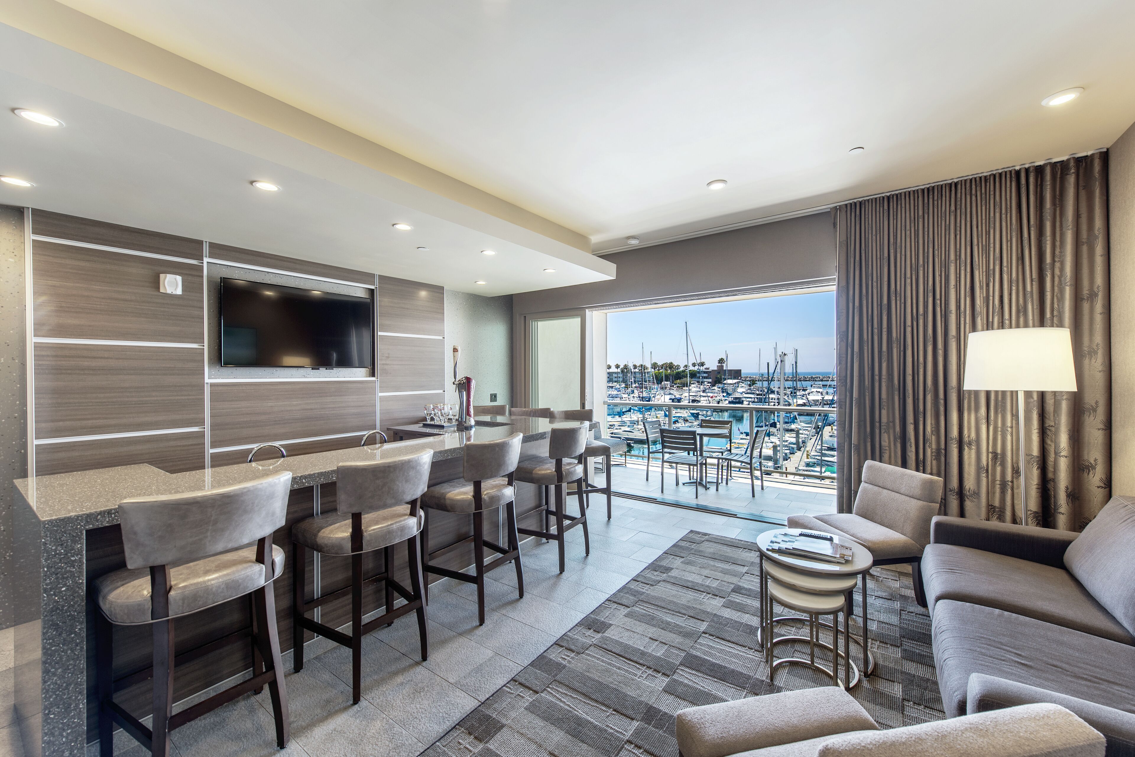 A modern hotel room with a bar area, seating, and a large window offering a marina view.