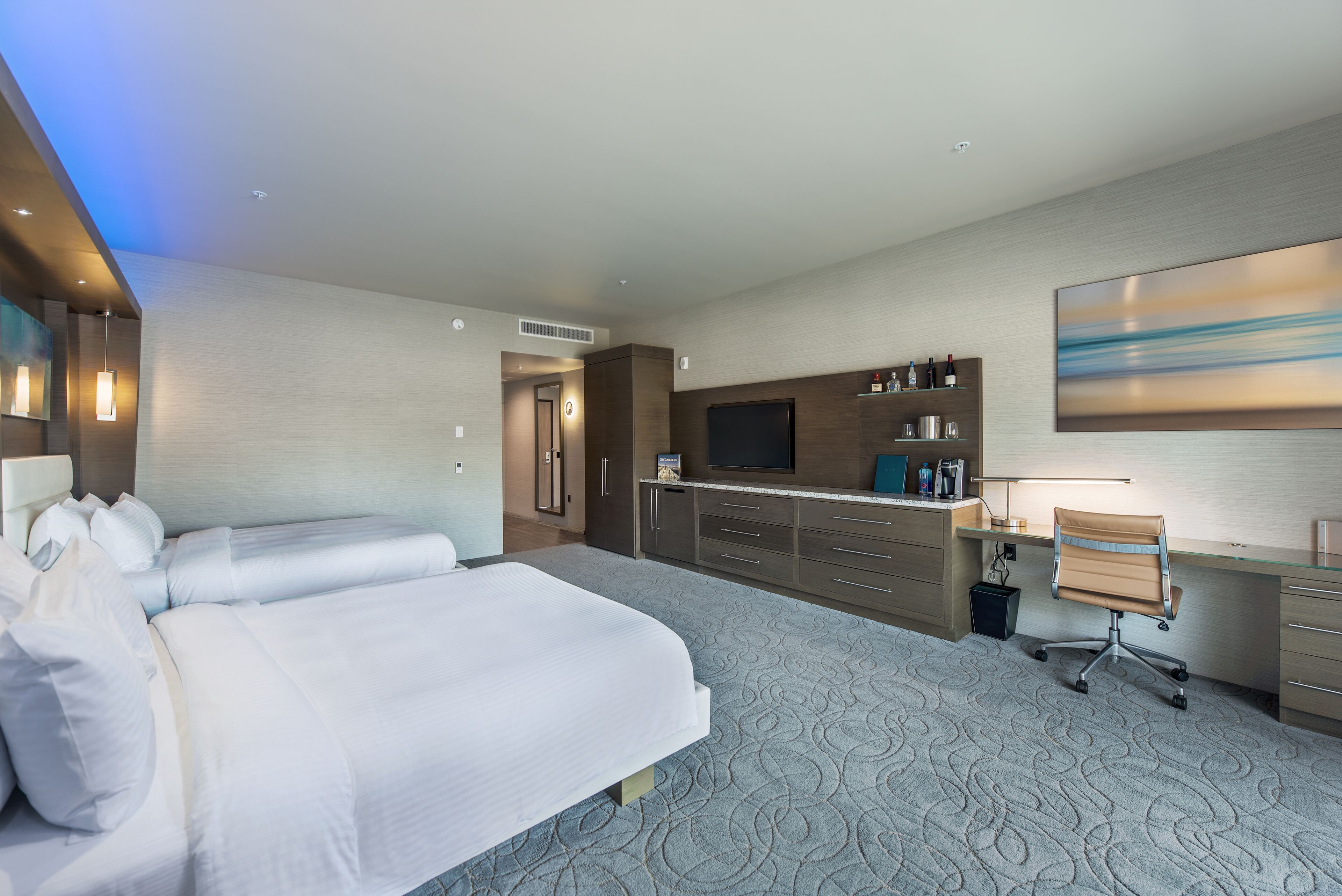 A hotel room with two beds, a desk, a chair, a TV, and a large abstract painting on the wall.