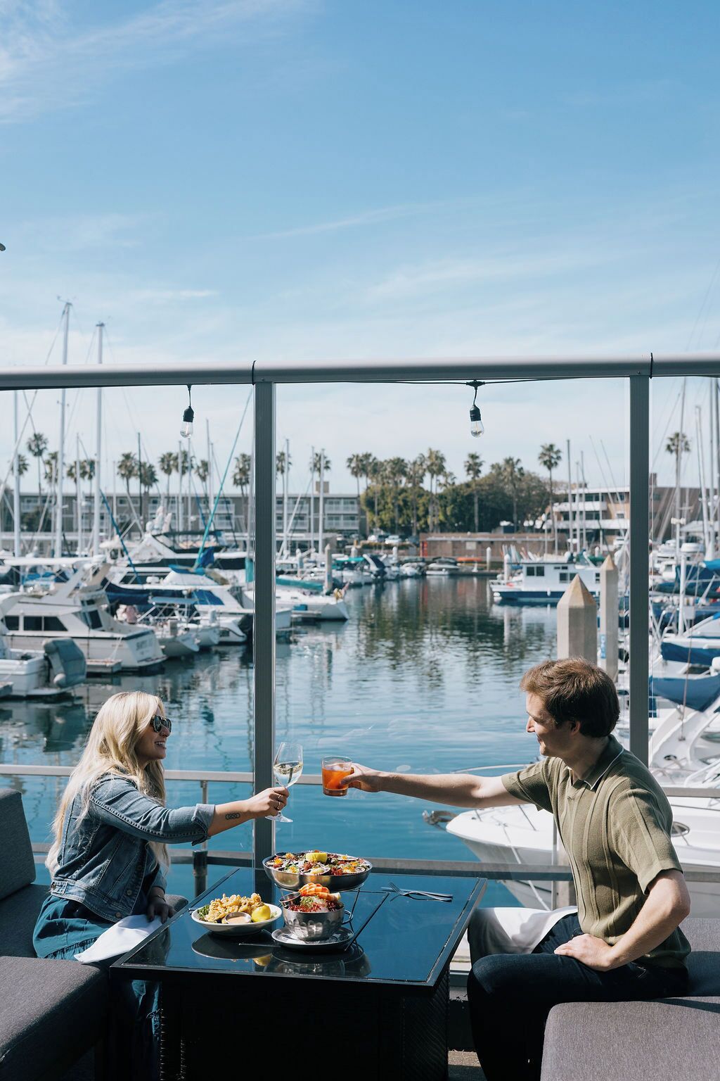 Un espace repas offrant une vue sur un port de plaisance, avec une table dressée avec de la nourriture et des boissons, et deux personnes qui trinquent.