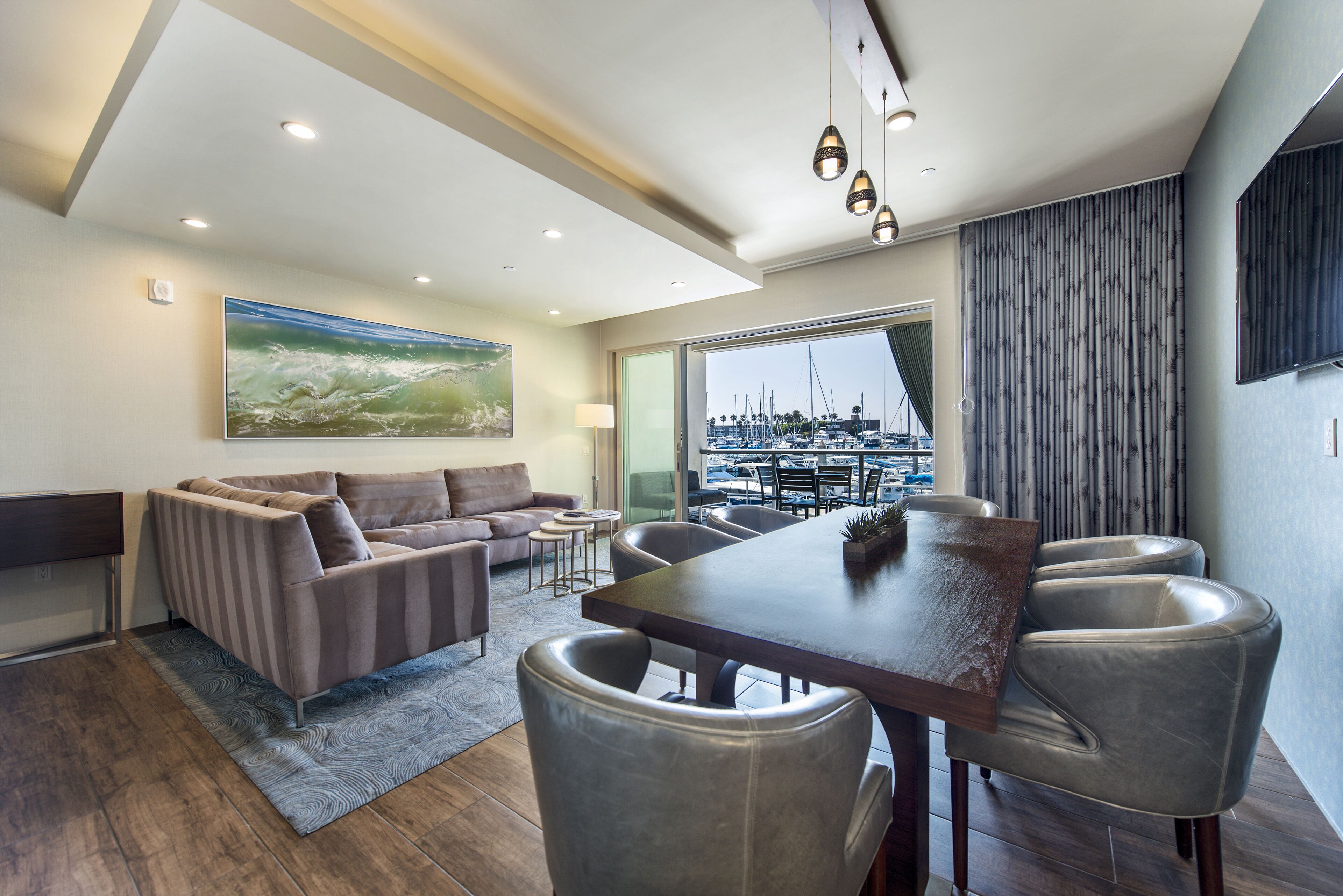 A modern living room with a sofa, dining table, and chairs, a large painting on the wall, and a view of a marina through a large window.
