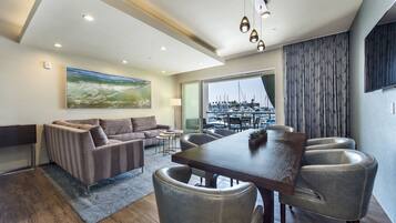 A modern living room with a sofa, dining table, and chairs, a large painting on the wall, and a view of a marina through a large window.
