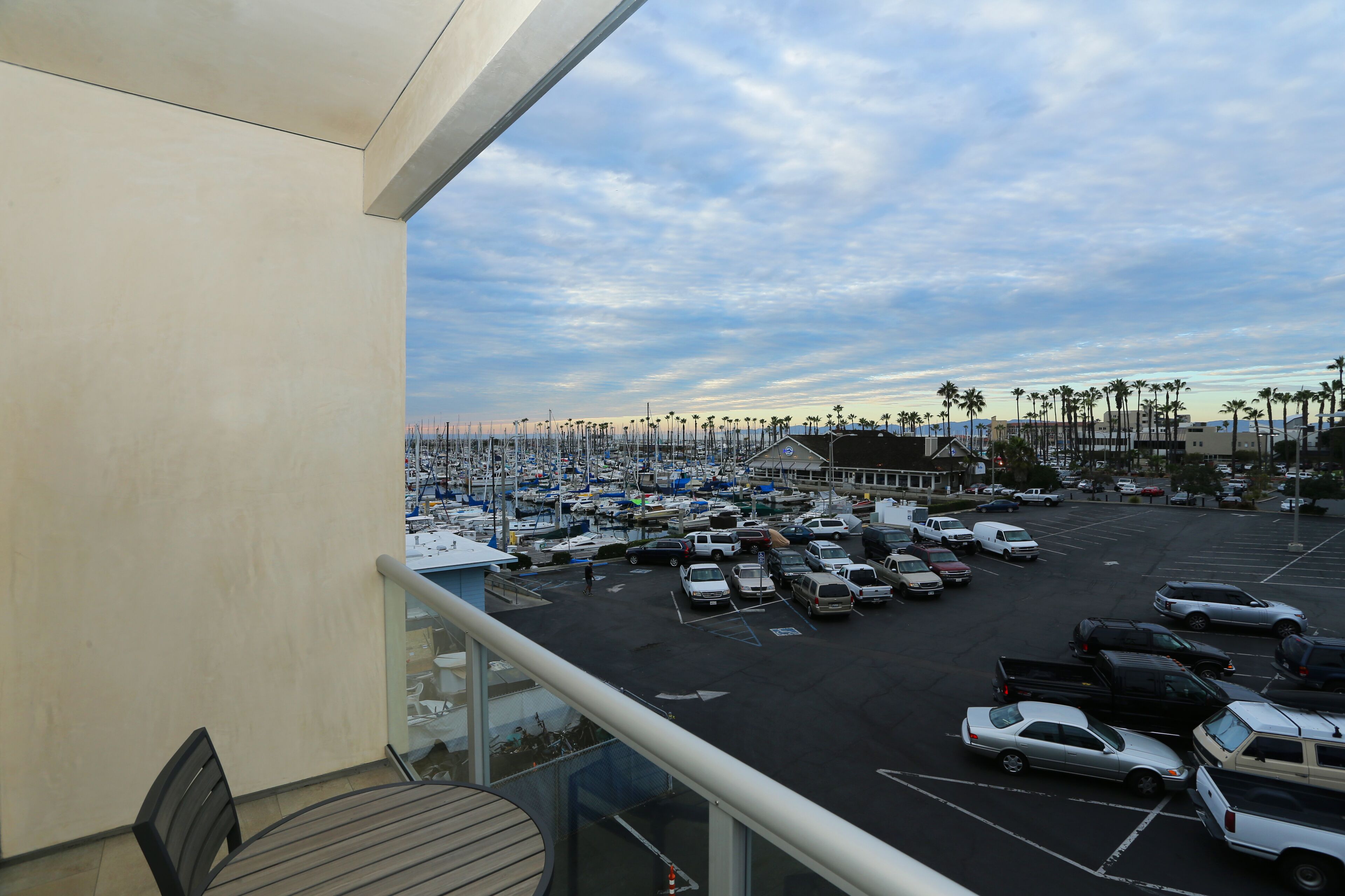 Un estacionamiento con varios vehículos, un puerto deportivo con barcos y palmeras al fondo.