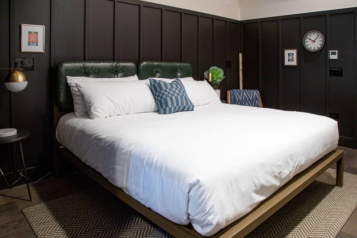 A modern bedroom with a large bed, dark wooden paneling, a clock, and a small potted plant.