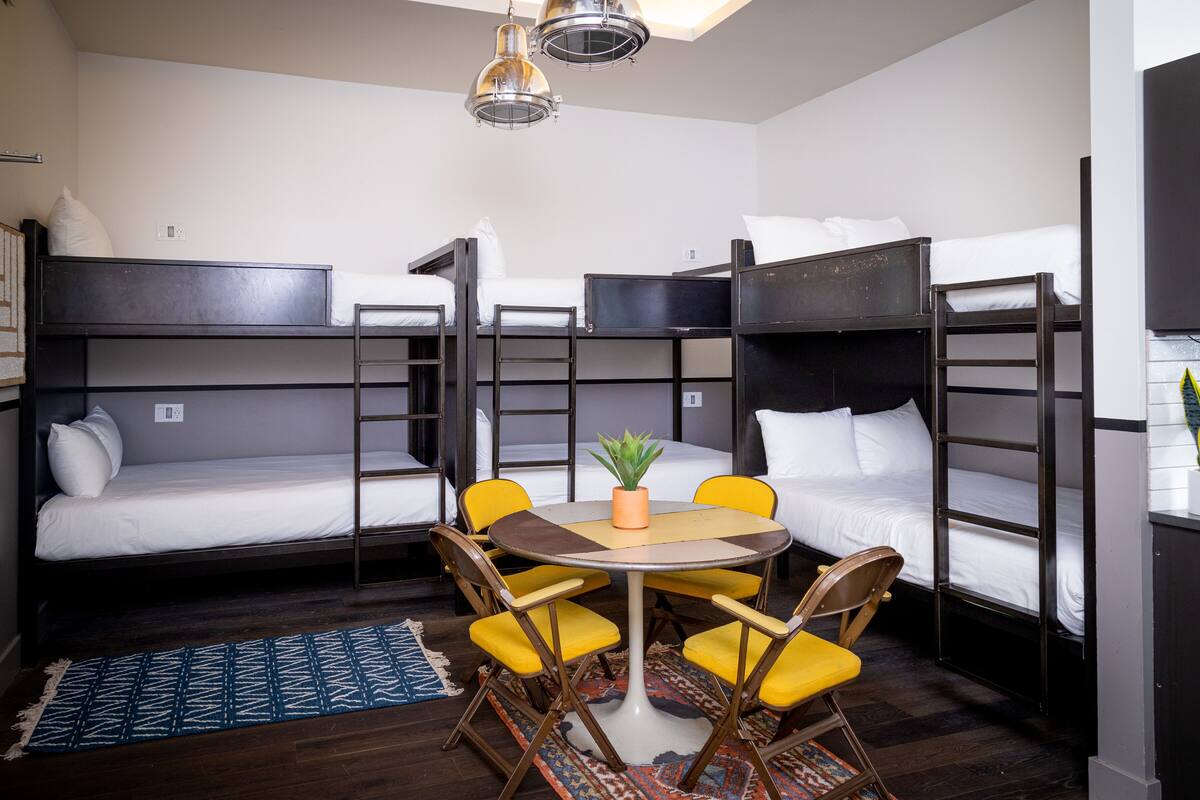 A dormitory room with bunk beds, a round table with chairs, and a small plant on the table.