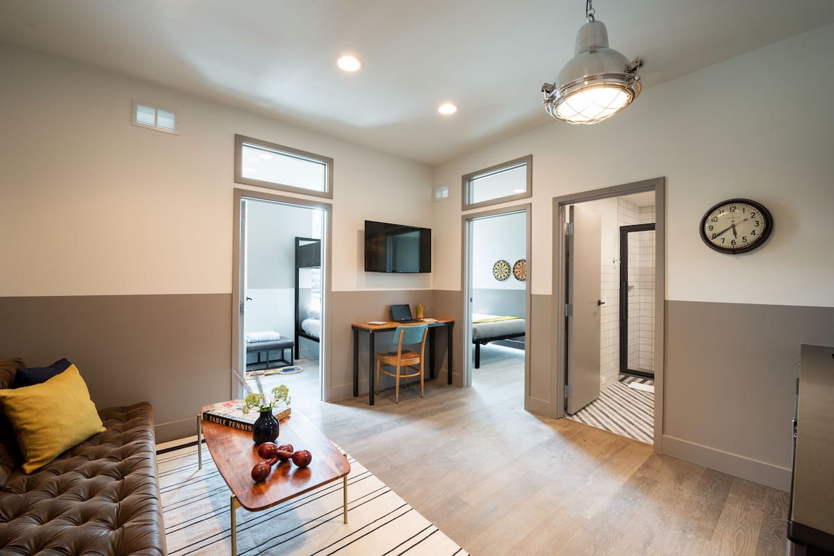 A modern living room with a sofa, coffee table, and a TV mounted on the wall. There is a dining area with a table and chairs, and a hallway leading to another room.