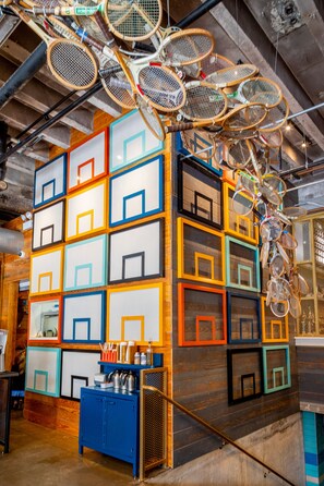 A room with a wall of colorful, framed geometric shapes, a blue cabinet with kitchen items, and a ceiling with hanging tennis rackets. - The Chicago Hotel Collection River North (Chicago)