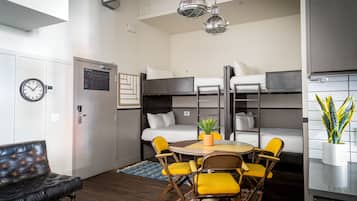 Habitación de hotel compacta con literas, una mesa de comedor con sillas amarillas, un sofá de cuero negro y una planta en maceta.