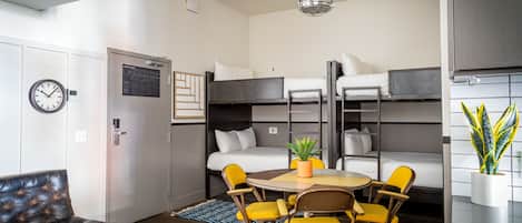 A compact hotel room with bunk beds, a dining table with yellow chairs, a black leather sofa, and a potted plant.
