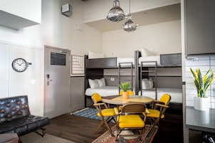 A compact hotel room with bunk beds, a dining table with yellow chairs, a black leather sofa, and a potted plant.