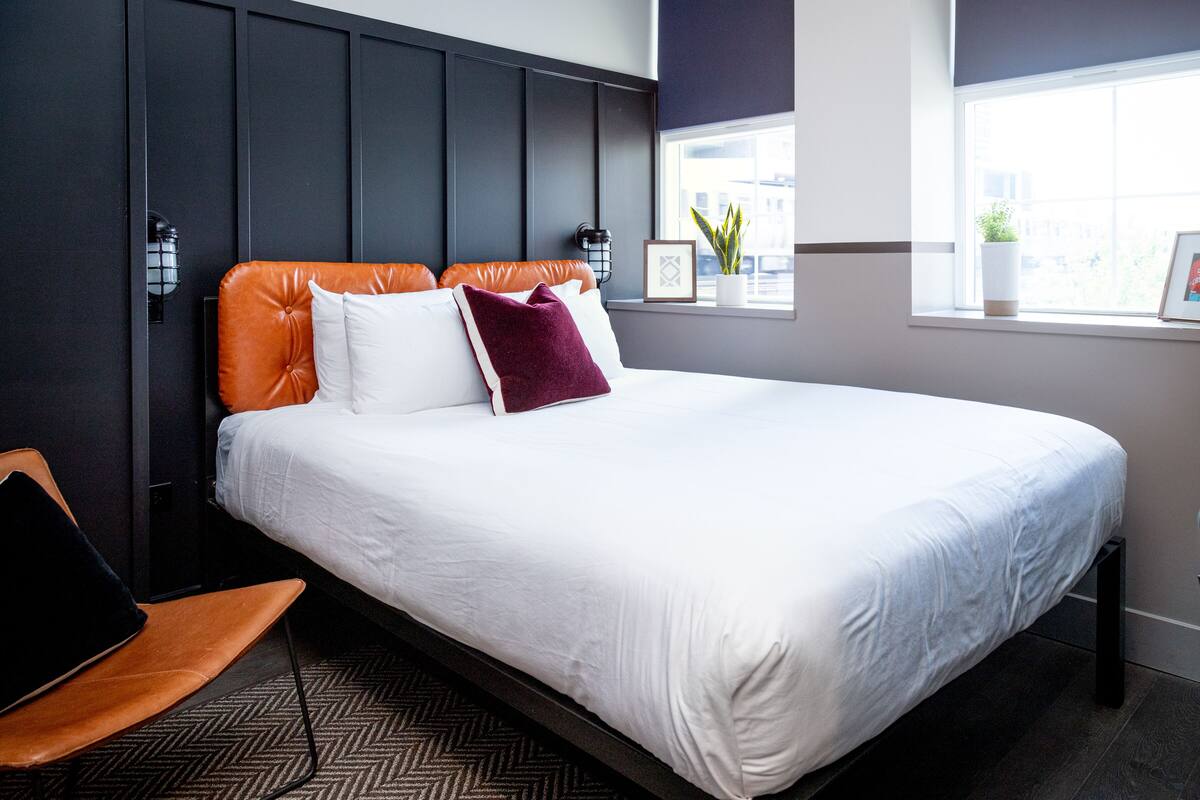 A neatly made bed with white linens, an orange headboard, and a purple pillow. There is a small potted plant on the windowsill and a framed picture on the wall.