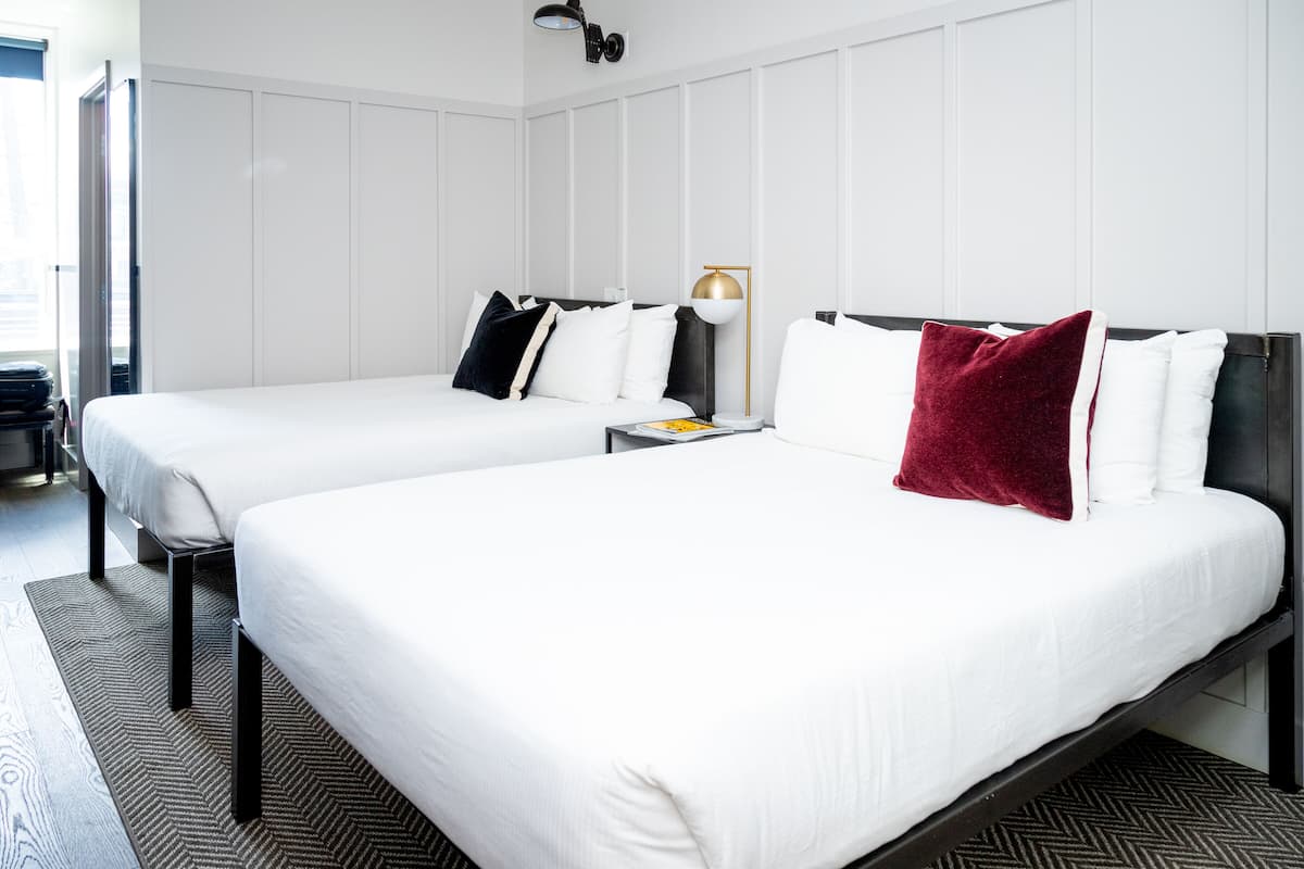 A hotel room with two beds, a nightstand, a lamp, and a large window.