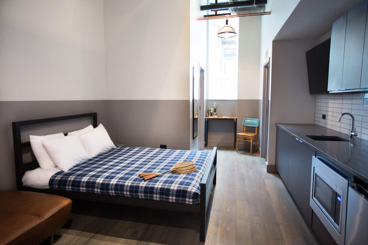 A compact hotel room with a bed, a small seating area, a kitchenette, and a bathroom.