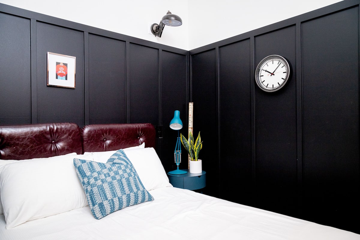 A bedroom with a dark wall, a clock, a framed picture, a bed with white and blue bedding, and a blue lamp.