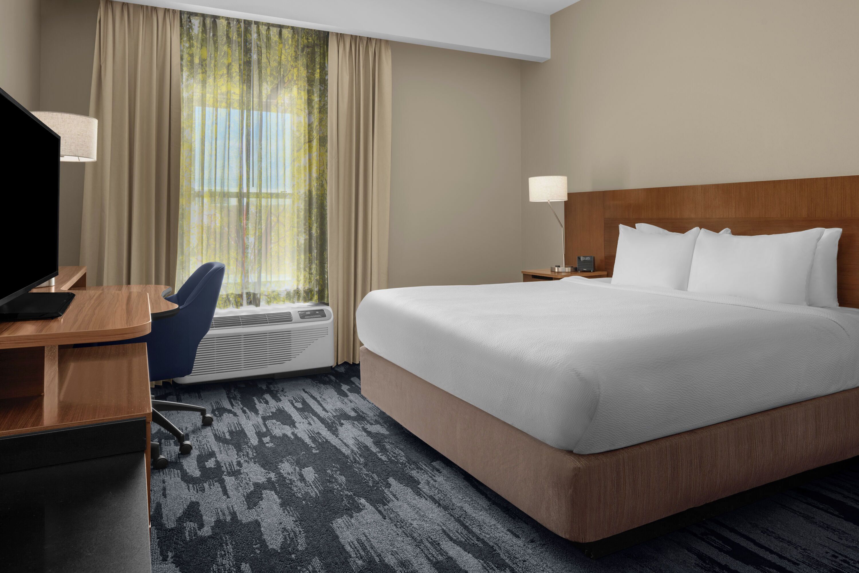 A hotel room with a large bed, a desk with a chair, a TV, a lamp, and a window with curtains.