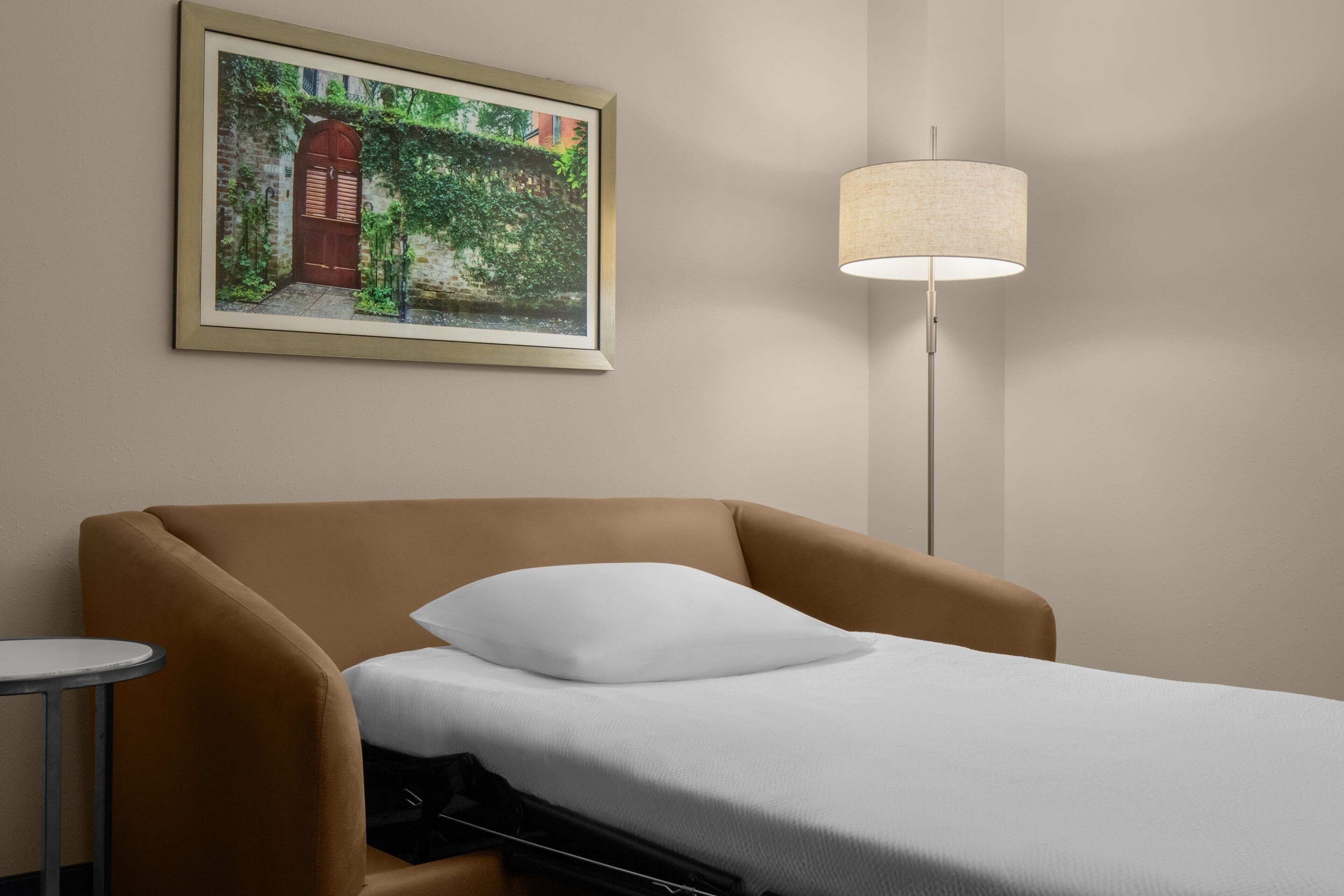 A hotel room with a bed, a small round table, a lamp, and a framed picture on the wall.