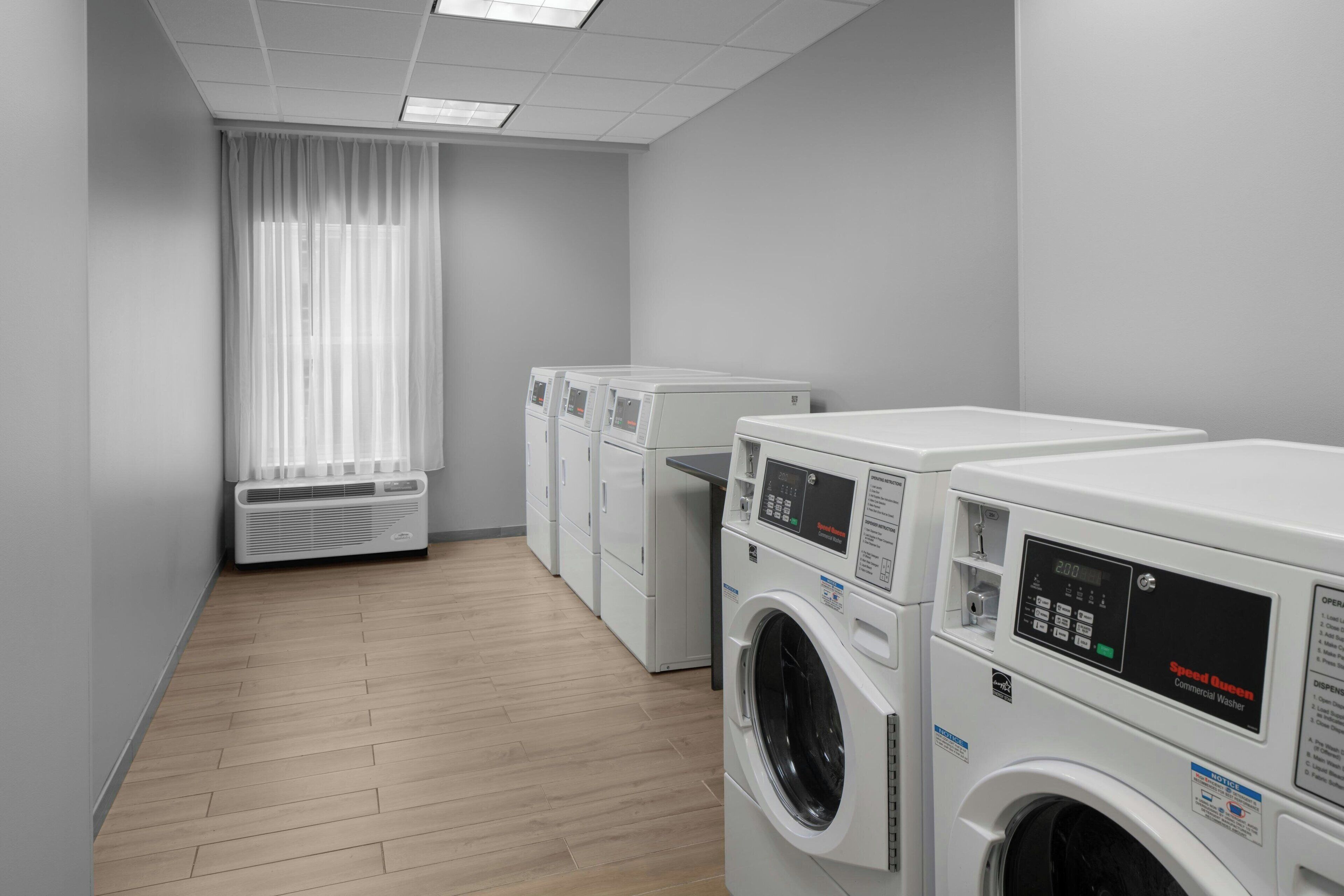 A laundry room with multiple commercial washers and dryers, a vented air conditioning unit, and a window with sheer curtains.