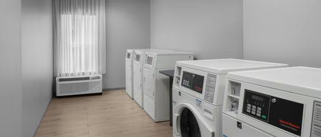 A laundry room with multiple commercial washers and dryers, a vented air conditioning unit, and a window with sheer curtains.
