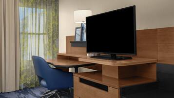 A hotel room with a wooden desk, a television, a blue chair, a lamp, and a window with curtains.