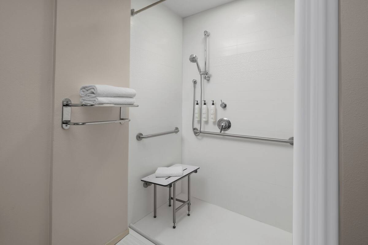 Un baño blanco con ducha, barras de apoyo, una mesita con una toalla y un estante con toallas.