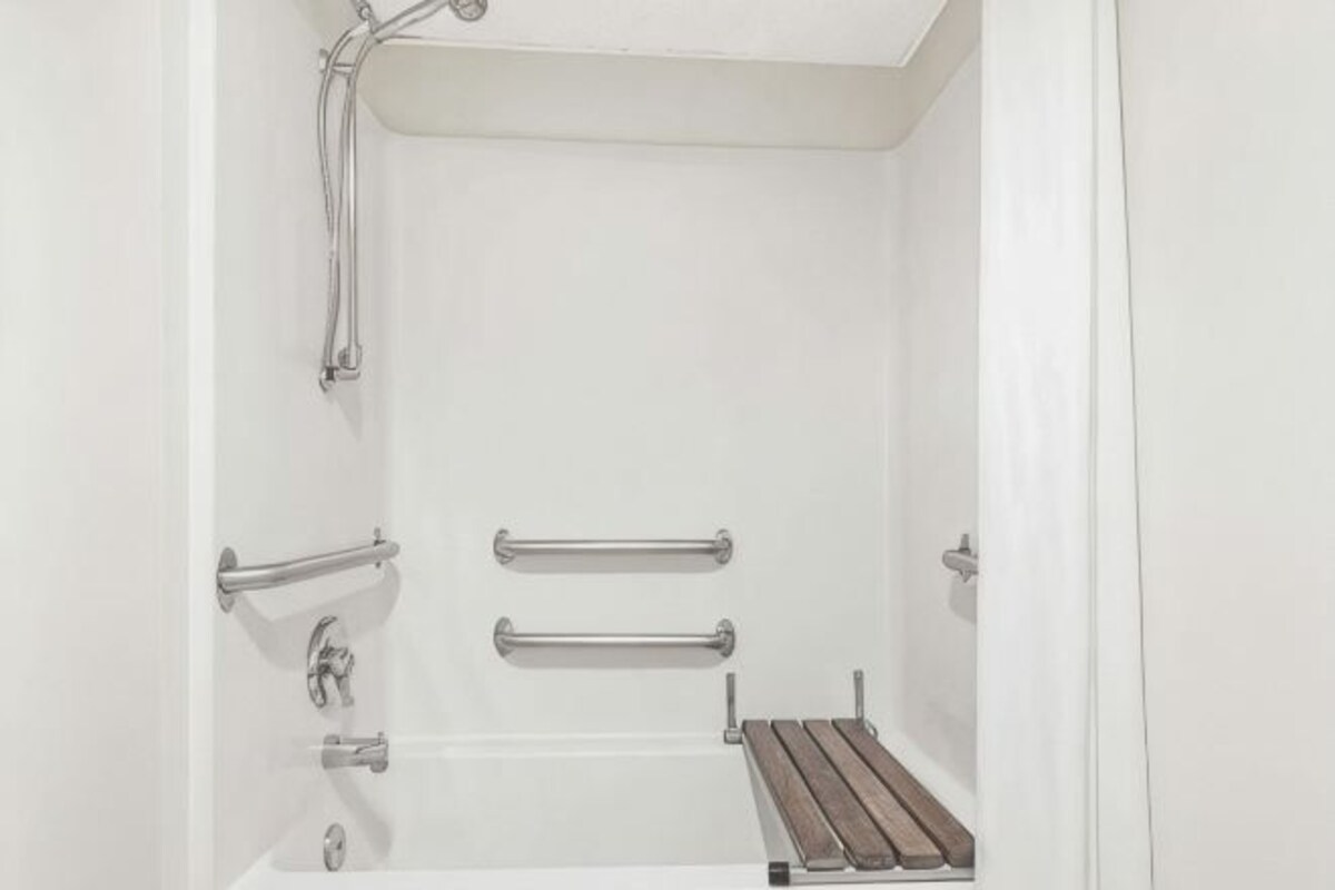 Un baño blanco con ducha, barras de apoyo y un estante de madera.