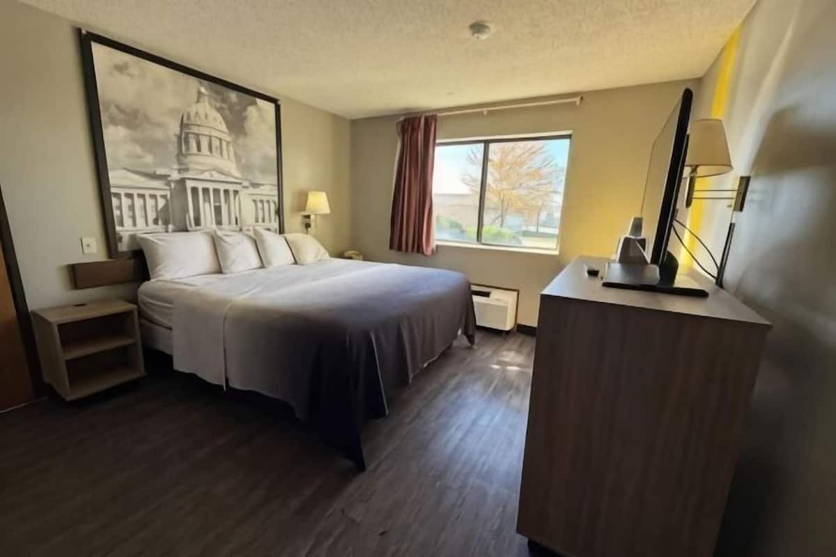 Habitación de hotel con una cama grande, un escritorio, una lámpara, una ventana con cortinas y un cuadro de un edificio.