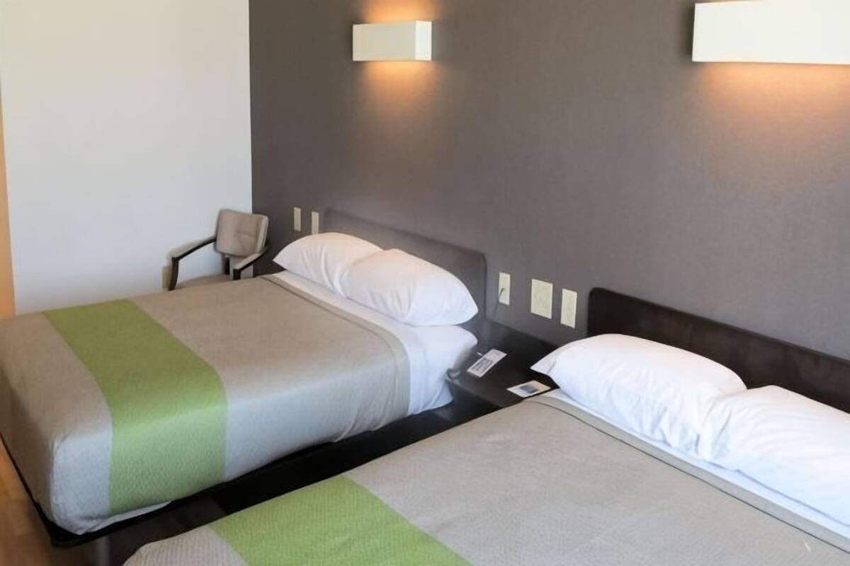 Habitación de hotel con dos camas, una silla, una mesita de noche e iluminación en la pared.