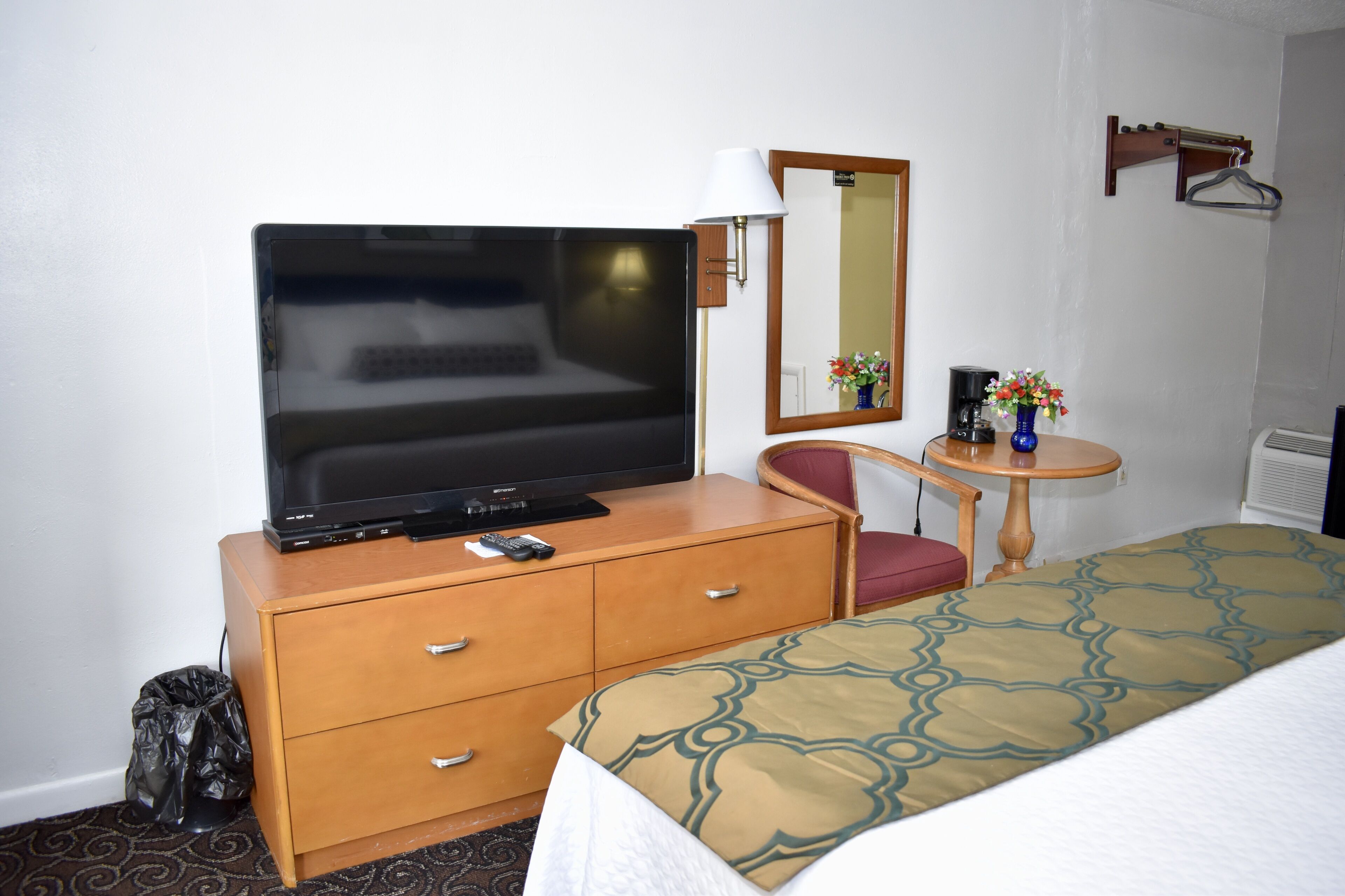 A hotel room with a bed, a television on a wooden dresser, a chair, a small table with a lamp, and a mirror on the wall.