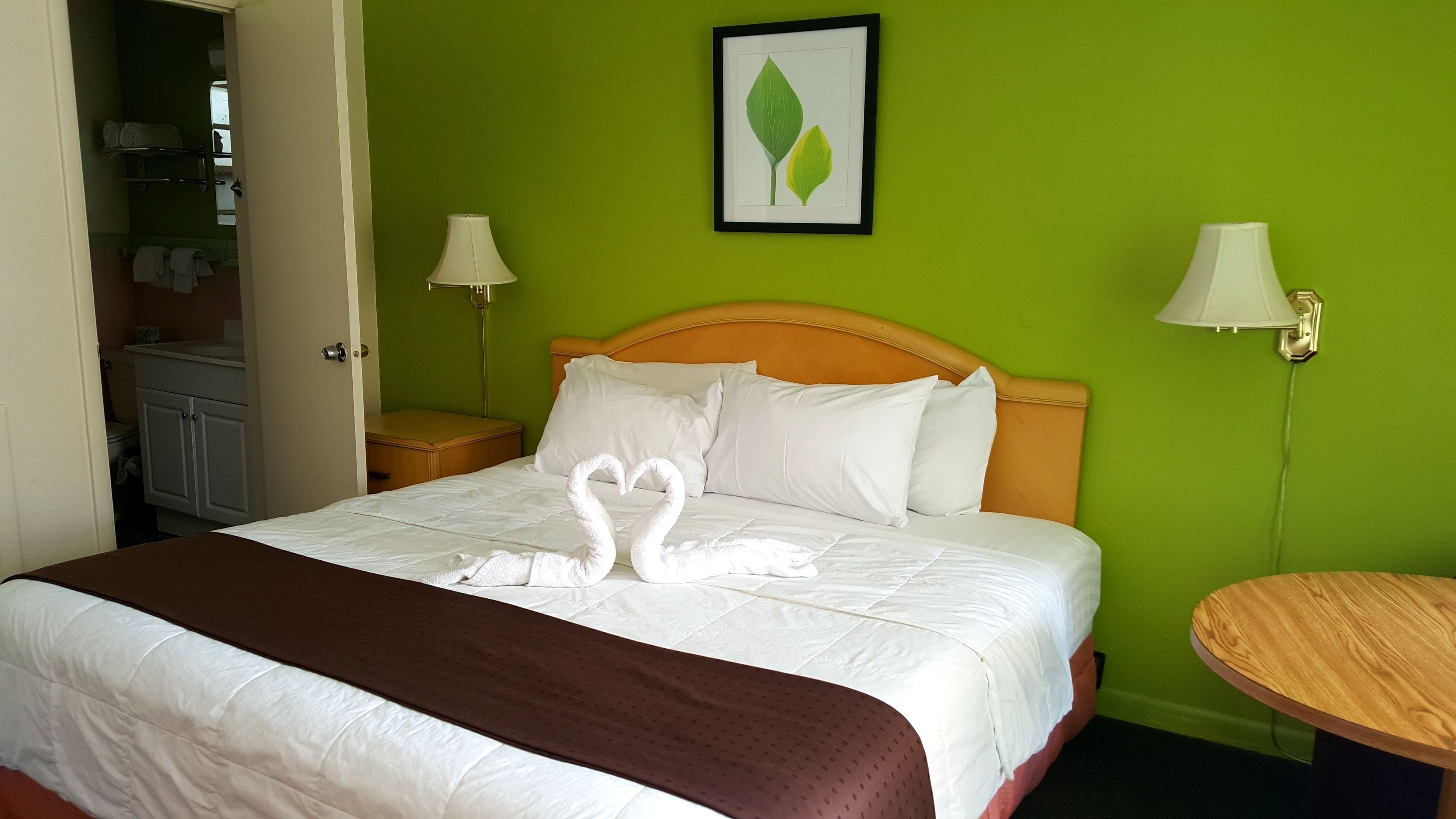 Une chambre d’hôtel avec un lit, une tête de lit en bois, deux tables de chevet avec des lampes, un mur vert et un tableau encadré au-dessus du lit.