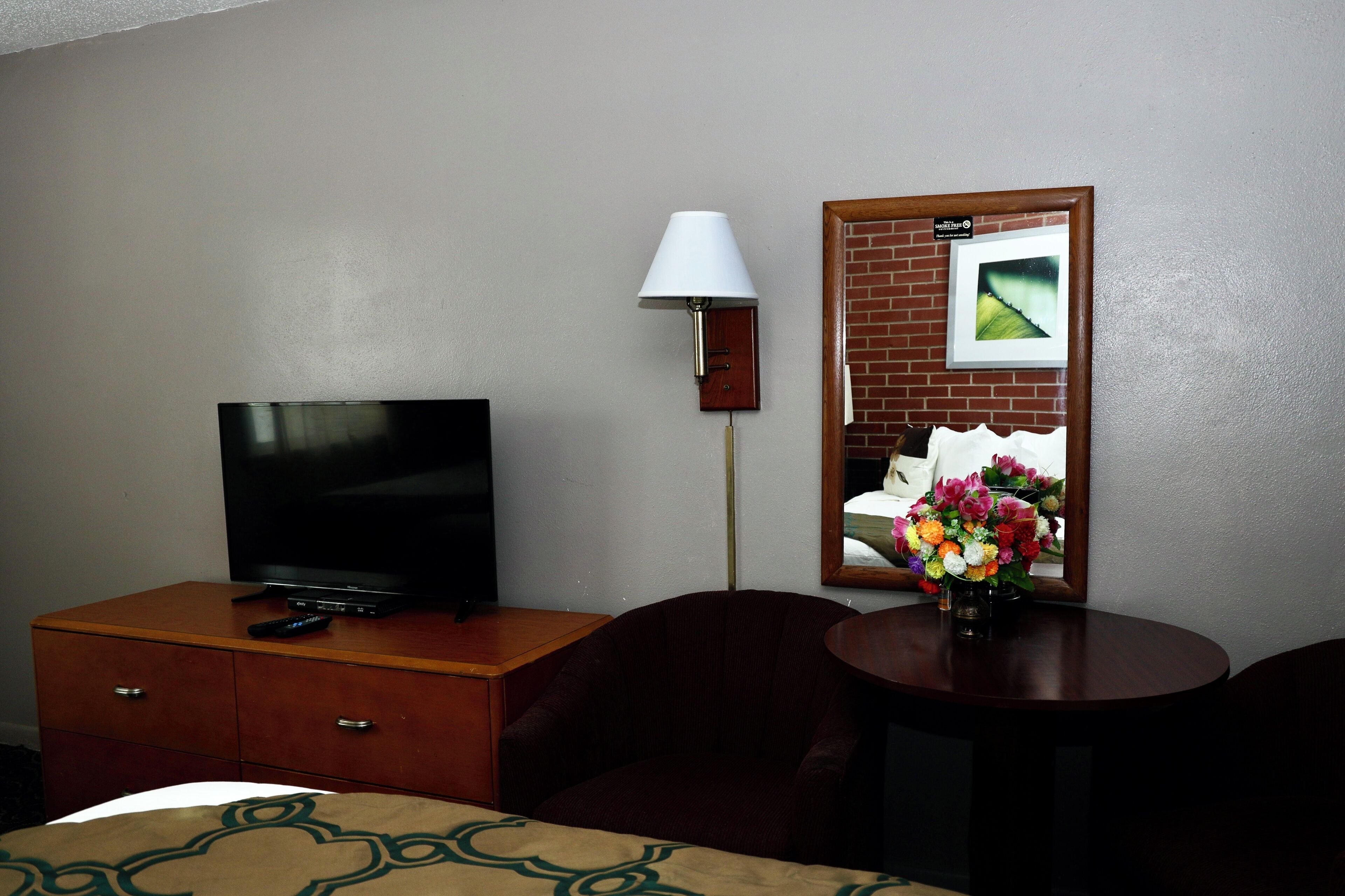 A hotel room with a TV on a wooden dresser, a lamp, a mirror with a picture, and a small table with a vase of flowers.