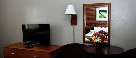 A hotel room with a TV on a wooden dresser, a lamp, a mirror with a picture, and a small table with a vase of flowers.