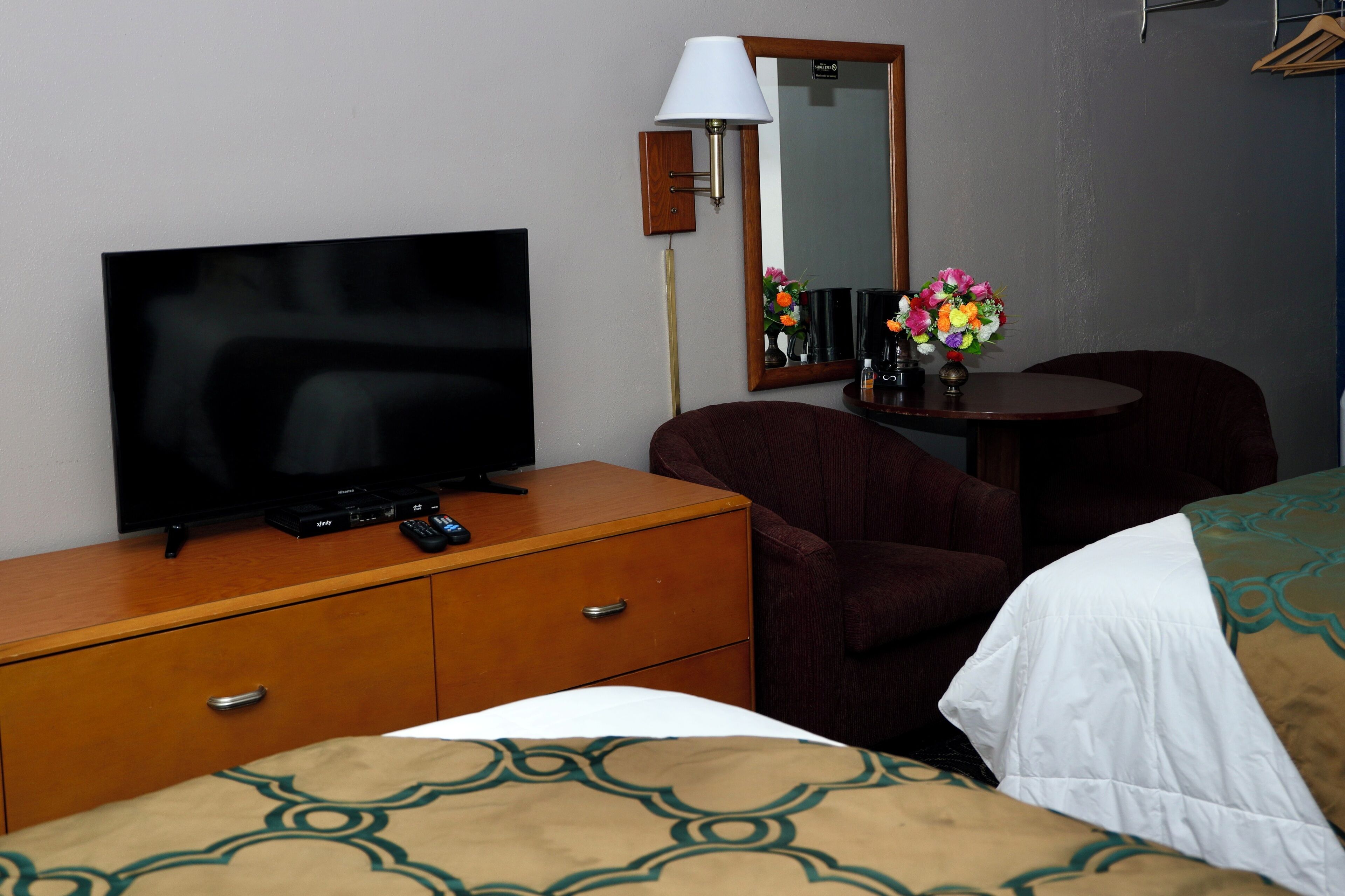 Ein Hotelzimmer mit Fernseher auf einer hölzernen Kommode, einem Spiegel, einem kleinen Tisch mit Blumenvase, einem braunen Sessel und einem Bett mit gemusterten Betttextilien.
