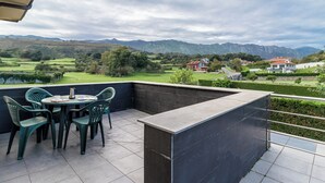 Une terrasse avec une table et des chaises, un mur en pierre et une vue sur un paysage verdoyant, avec des maisons et des montagnes à l’horizon.