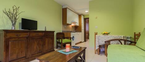 A living room with a wooden table, a sofa, a television, and a kitchen in the background.