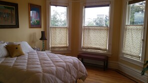 A bedroom with a large bed, two windows with patterned curtains, a bedside table with a lamp, and a bench. - Gorgeous 4 Bedroom Victorian Townhouse (Oakland)