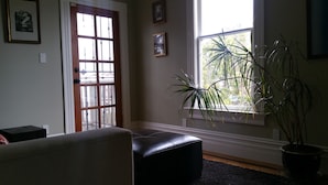 A living room with a black leather ottoman, a large window with a view of greenery, and a door with glass panels.