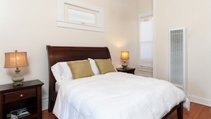 A bedroom with a large bed, two bedside tables, a wall-mounted lamp, a ceiling light, a window with blinds, and a radiator.