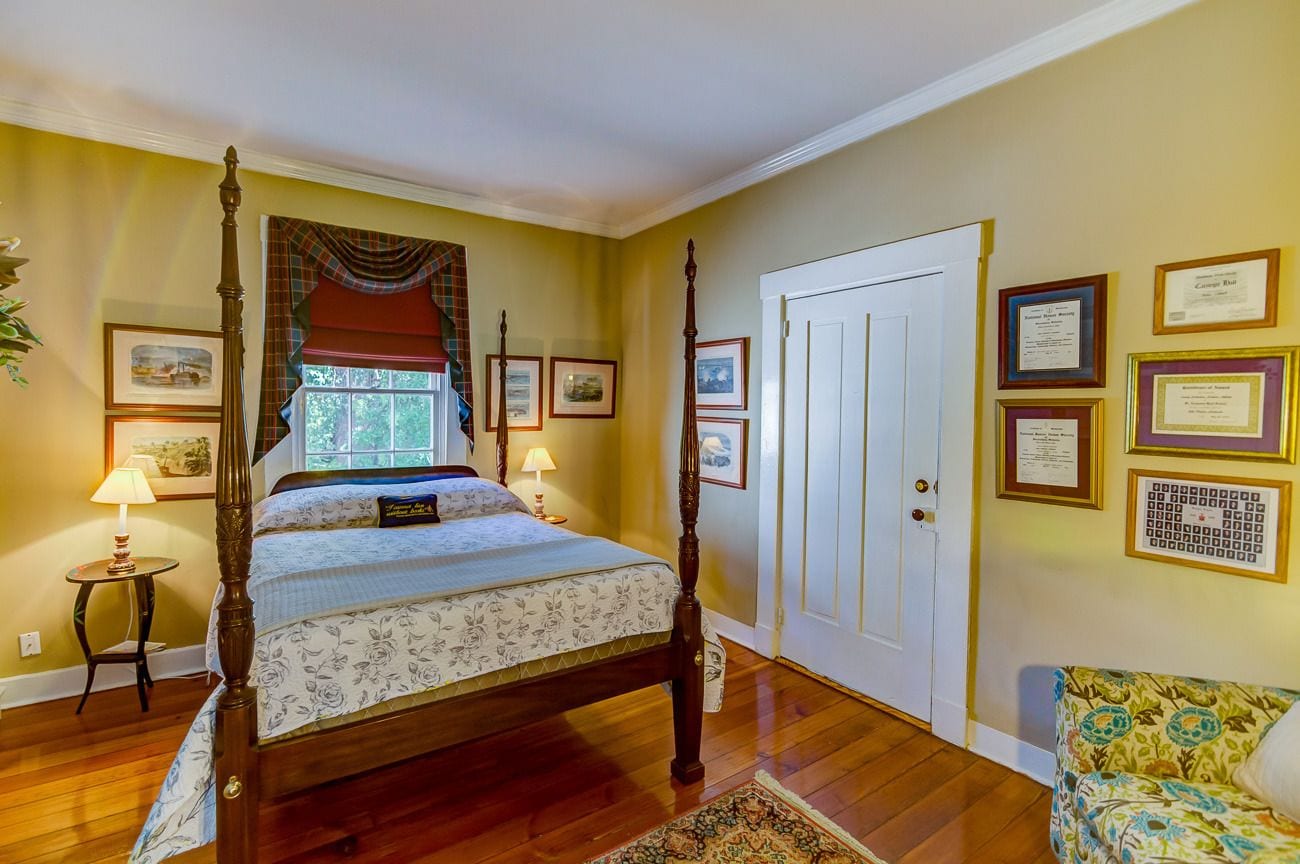 A bedroom with a four-poster bed, a window with curtains, a bedside table with a lamp, and a door with framed certificates on the wall.
