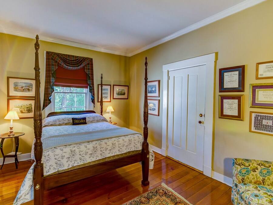 A bedroom with a four-poster bed, a window with curtains, a bedside table with a lamp, and a door with framed certificates on the wall.