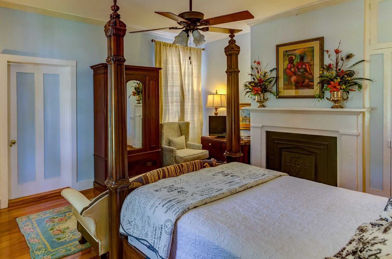 A bedroom with a four-poster bed, a wooden chair, a desk, a fireplace, and a ceiling fan.