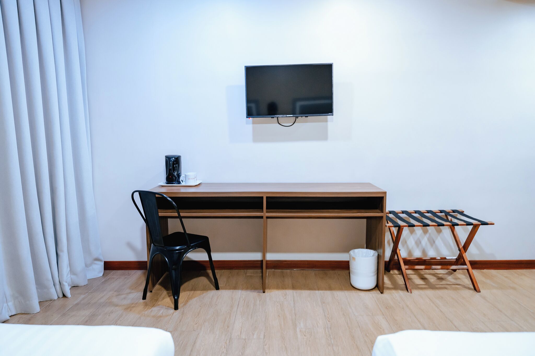 A room with a wooden desk, a black chair, a TV mounted on the wall, a small table with a cup, and a folding chair.