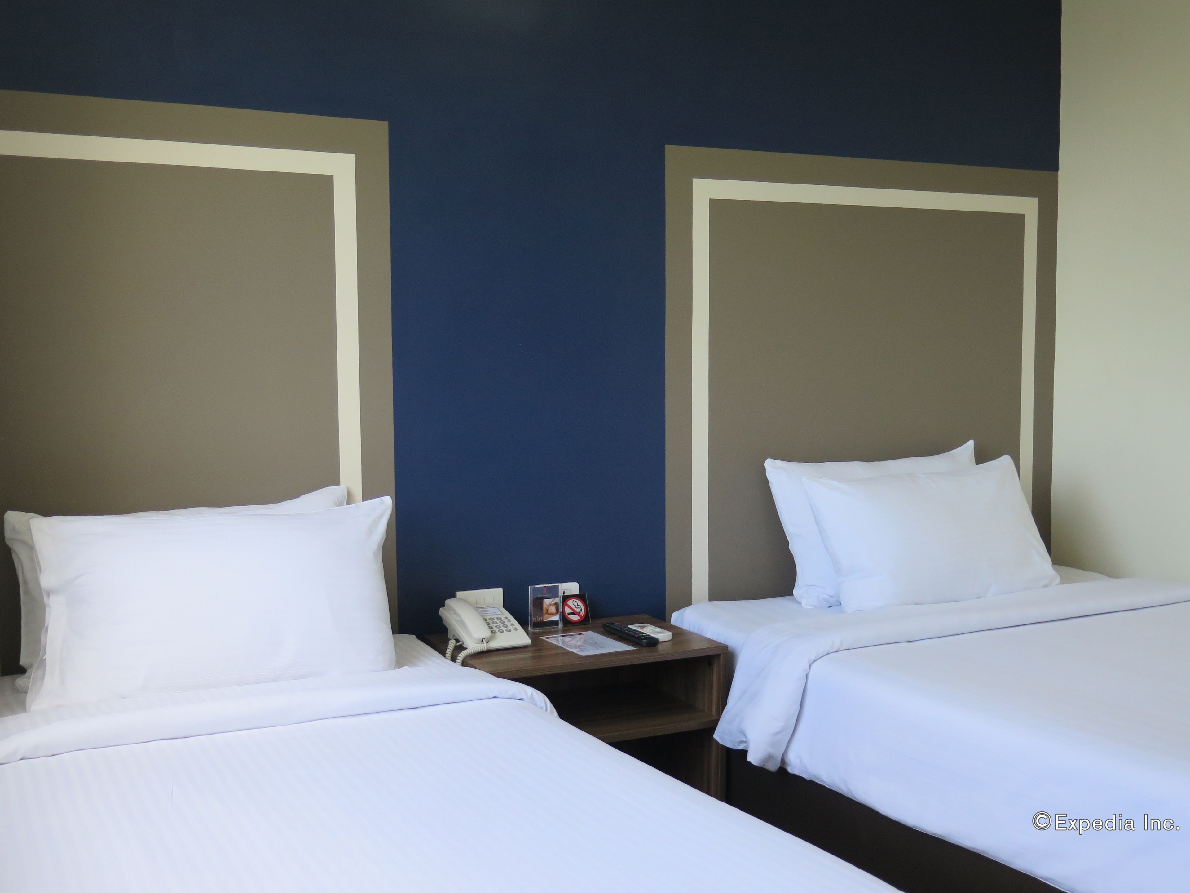 Une chambre d’hôtel avec deux lits, chacun équipé de draps blancs, une table de chevet avec un téléphone et une télécommande, et un mur orné d’un motif géométrique.
